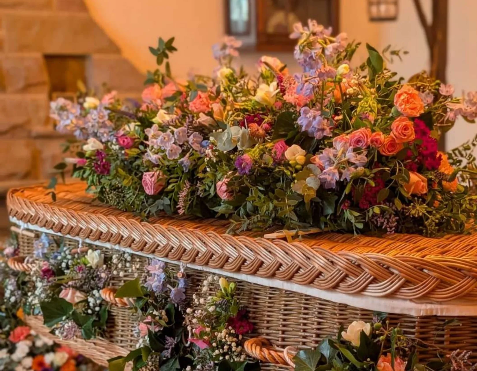 A meadow-style casket spray of pink, lilac and blue flowers on a fully decorated wicker coffin