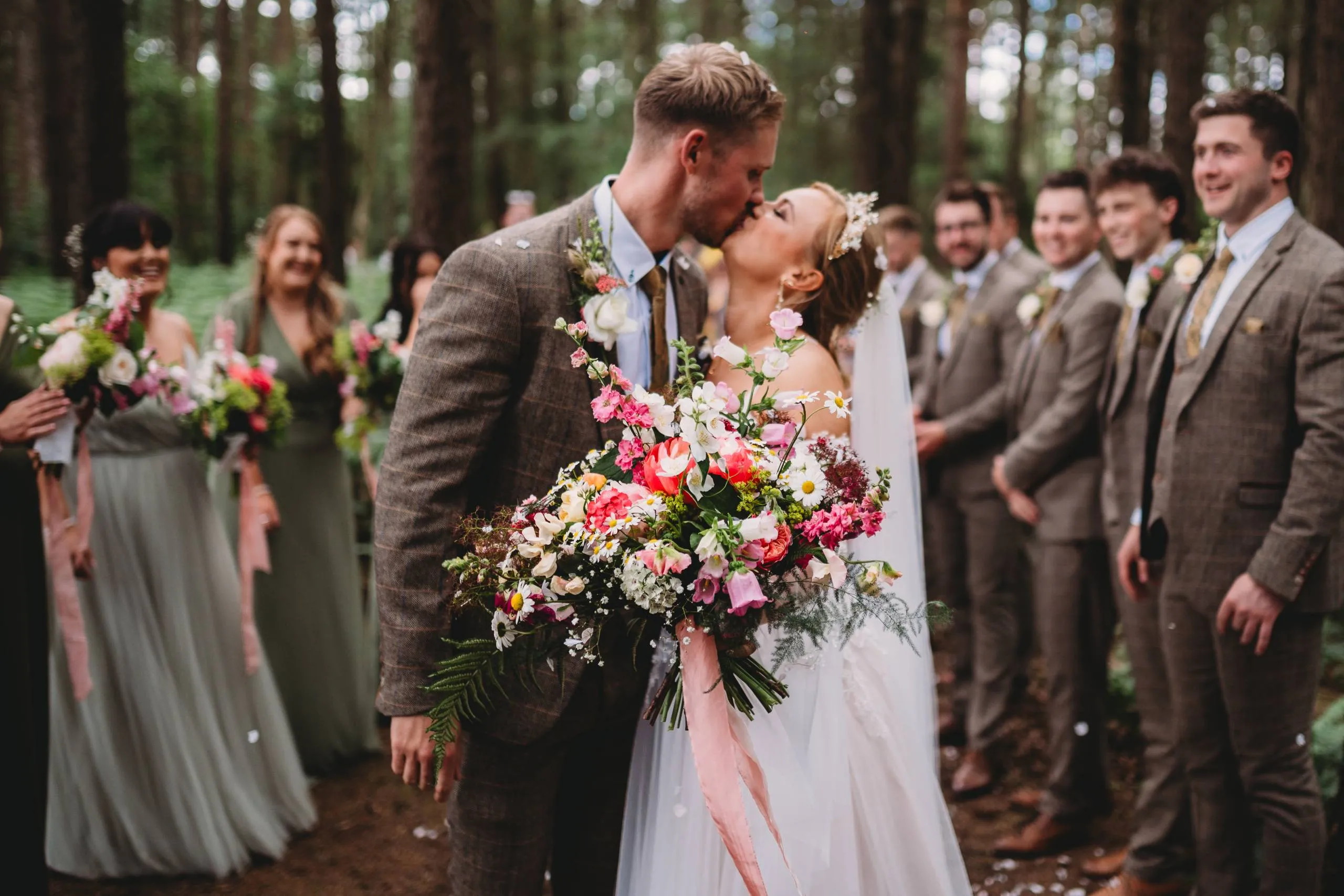 A couple kissing in woodland, surrounded by the bridal party, with a wild seasonal bouquet