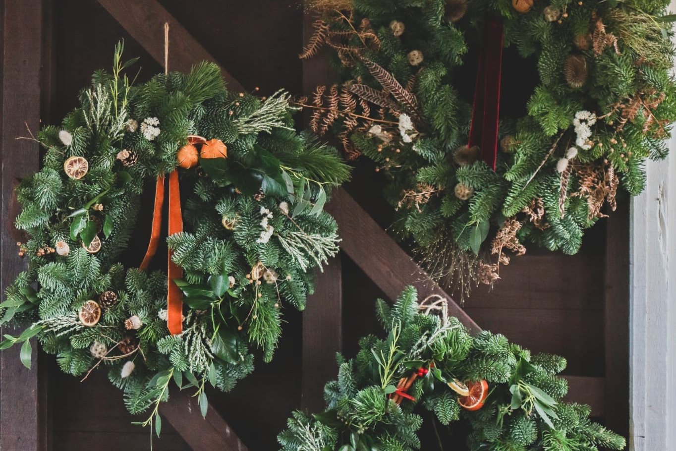 Three finished Christmas wreaths on a dark door, with dried orange slices and ribbon